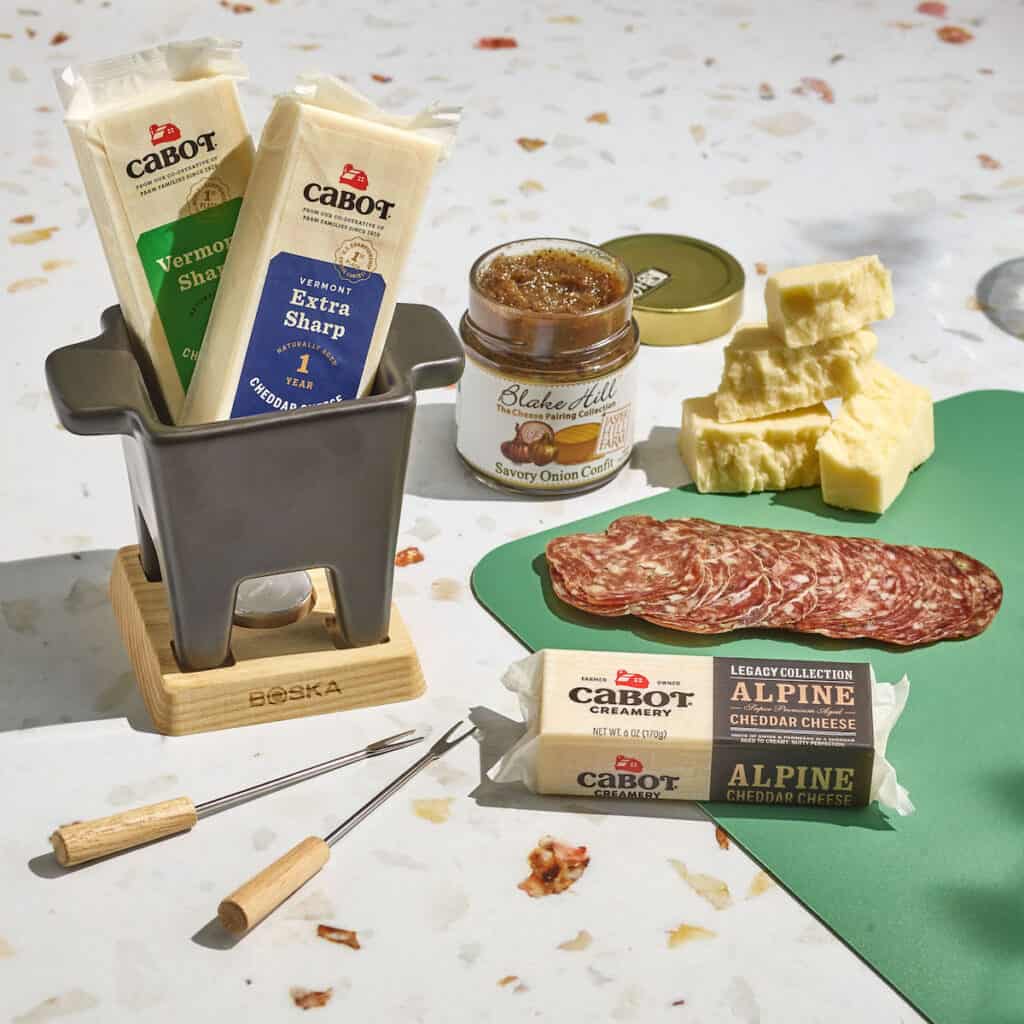 A spread of ingredients for fondue includes sharp cheddar cheeses, alpine cheddar, cured meats and savory onion confit.
