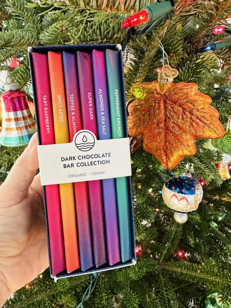 A box of full size dark chocolate bars showing the color "spines" of the bars as if they are a stack of books. A lit and decorated Christmas tree is the backdrop.