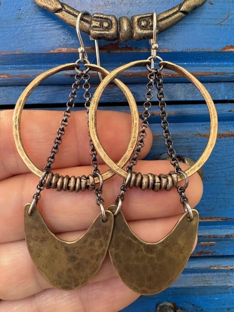 A set of bronze colored metal earrings hang from a blue wooden jewelry case. Fingers present the earrings to you.