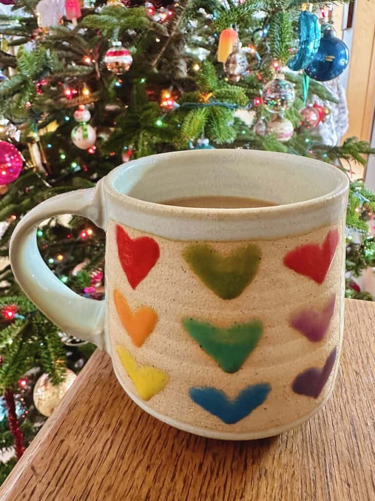 An off white mug with a rainbow palette of painted hearts on it sits in front of a lit and decorated Christmas Tree. It a perfect Vermont made gift that screams LOVE!
