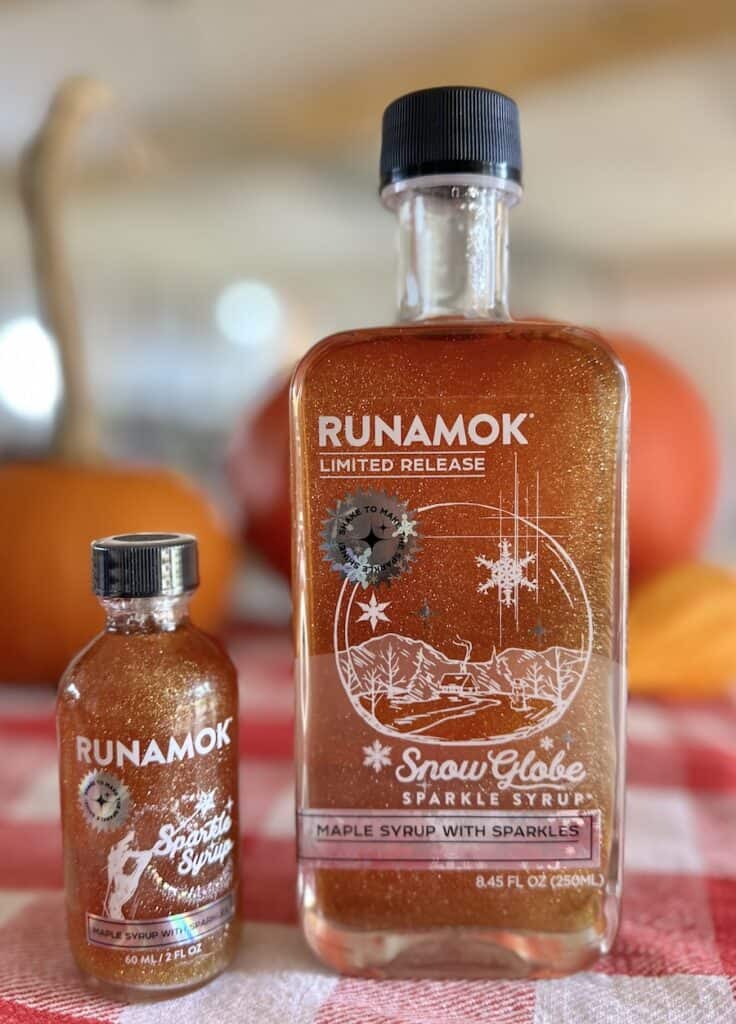A stocking size and a full size bottle of Runamok Snow Globe Sparkling maple syrup sit on a red and white checkered cloth. Orange pumpkins are in the background.