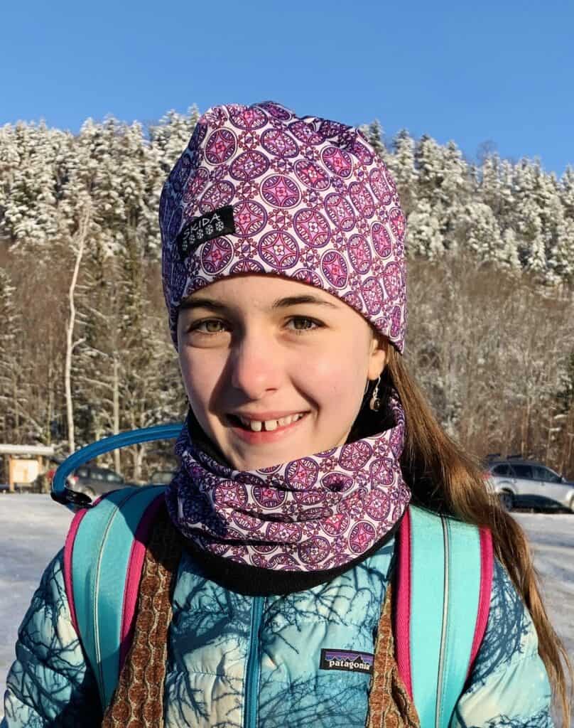 My daughter, Lucy, wearing a purple, pink, and white Skida hat and a neck buff with the same pattern. It's winter and she has a blue and white Patagnoia jacket on.