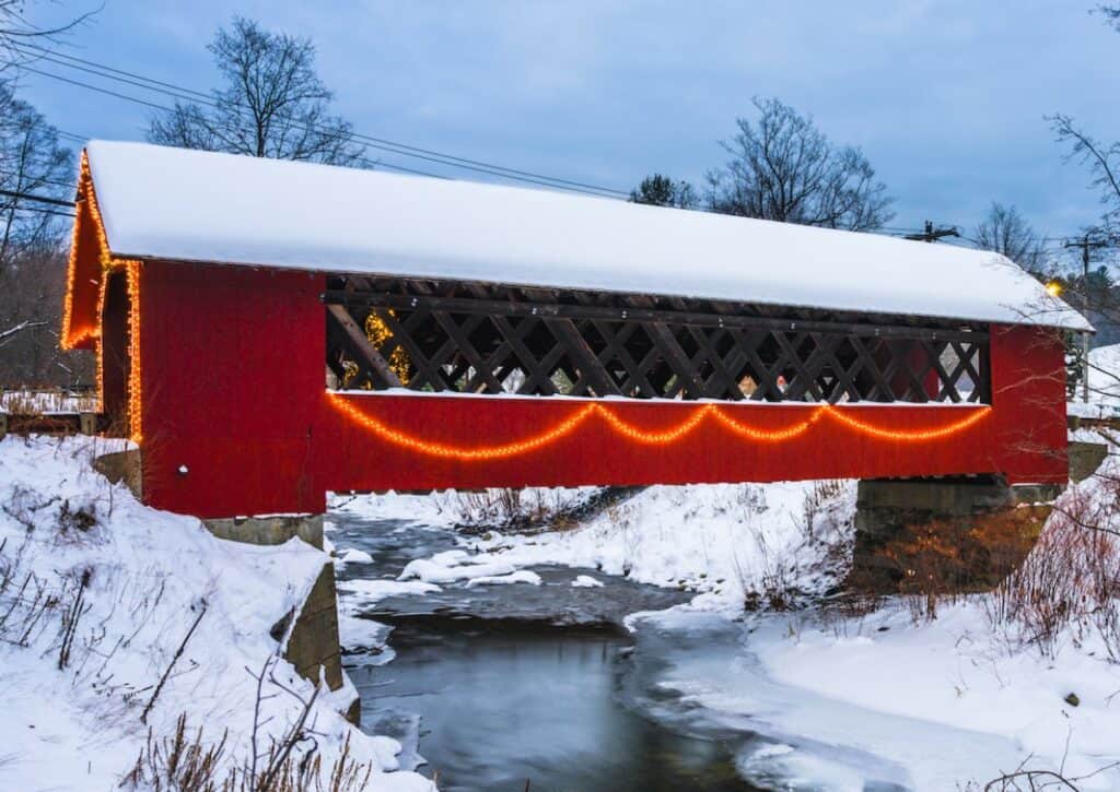 A red covered bridge in Vermont is decorated in a snowy winter scene with holiday lights.