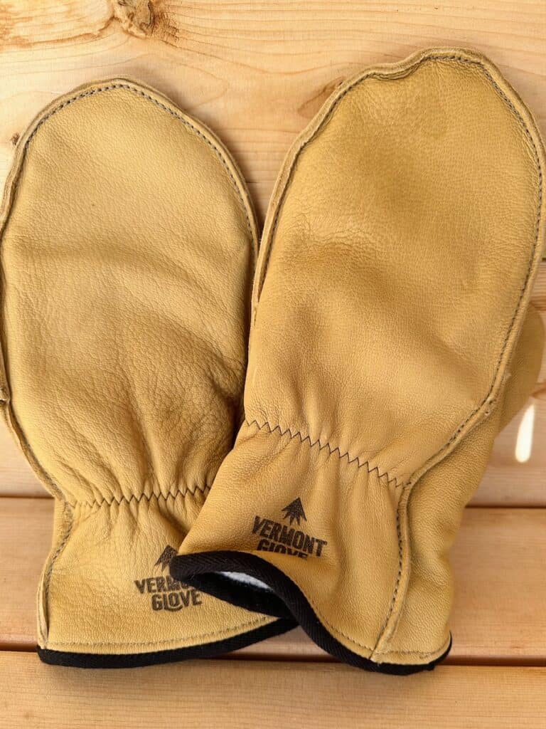 A pair of Vermont Glove made mittens rest on a wooden surface. They are golden tan and have a black edge on the cuff.