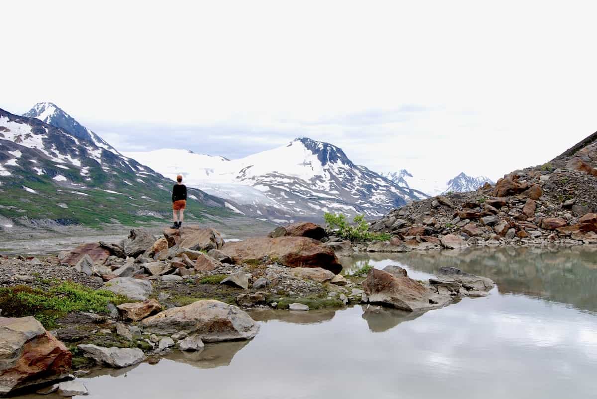 Here I am standing on a rocky outcrop at Iceberg Lake, surrounded by the tranquil beauty of snow-capped mountains and a partially frozen lake reflecting the alpine scenery in Alaska. The overcast sky adds a serene atmosphere to the wilderness landscape.