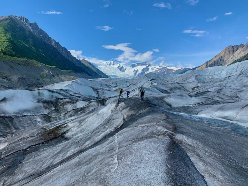 My husband, Matt, and my kids are equipped with backpacks as we traverse the rugged, crevassed surface of the Root Glacier in Alaska, with a dramatic view of the glacier flowing between steep mountains under a bright blue sky. The natural striations and melting patterns of the glacier's ice create a textured landscape leading towards the distant icefall.