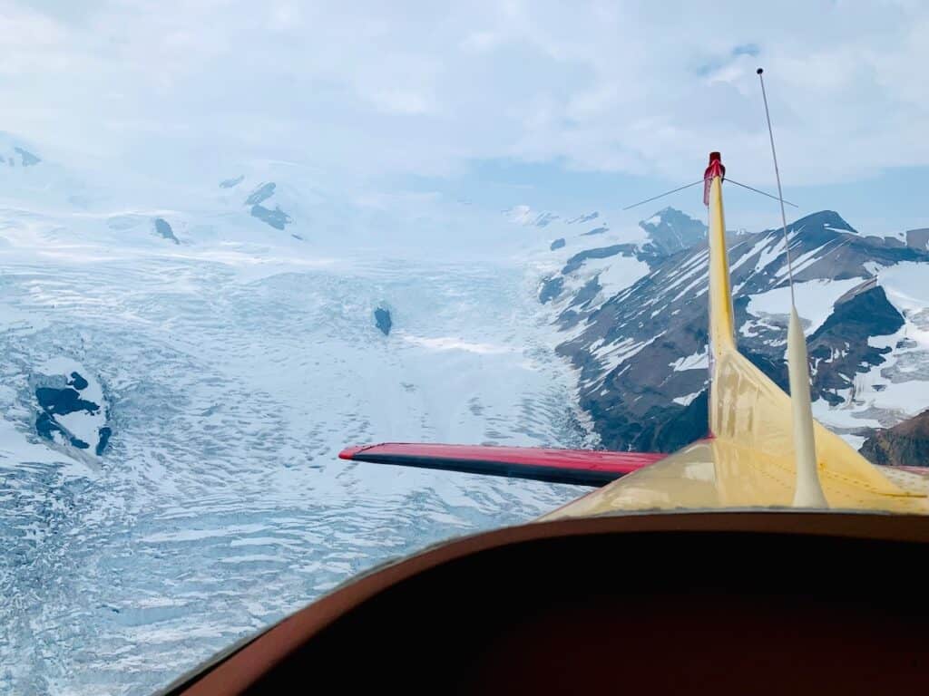 View from a small aircraft with a yellow nose, overlooking the vast, icy expanse of glaciers and rugged mountains in McCarthy, Alaska. The wing's edge and antenna are visible, pointing towards the scenic, snowy landscape below.